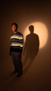 Toronto-based R&B artist Jon Vinyl wearing a strip shirt with a spotlight lighting his upper body in a dark almond tone room.